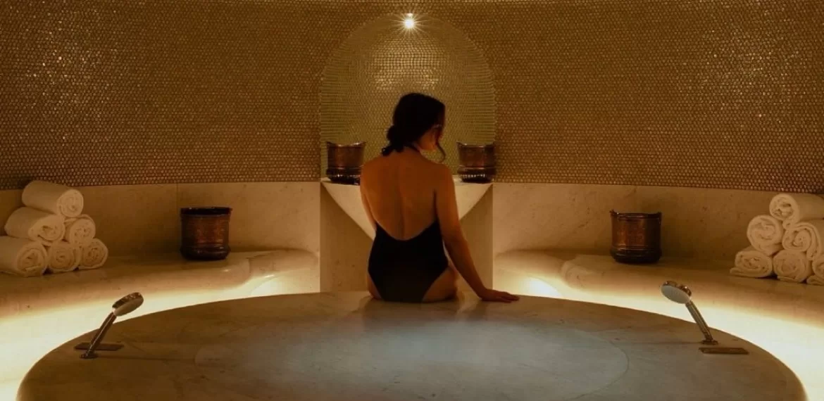 Woman sitting with her back to camera on a marble hammam platform beneath a gold mosaic dome, copper buckets and rolled towels either side
