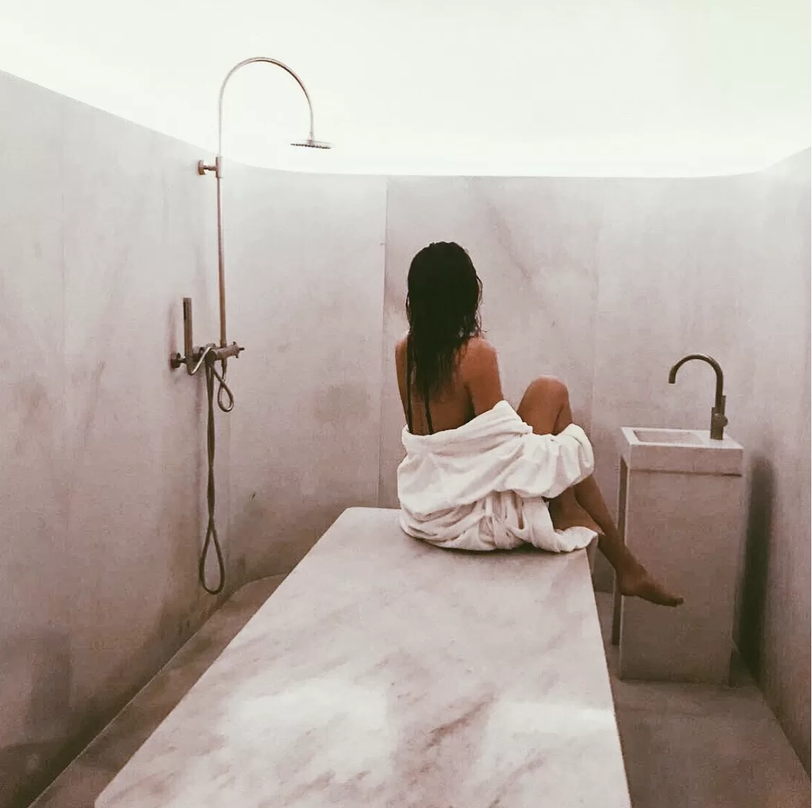 Woman wrapped in a white towel sitting on a marble slab in a minimalist hammam-style wet room