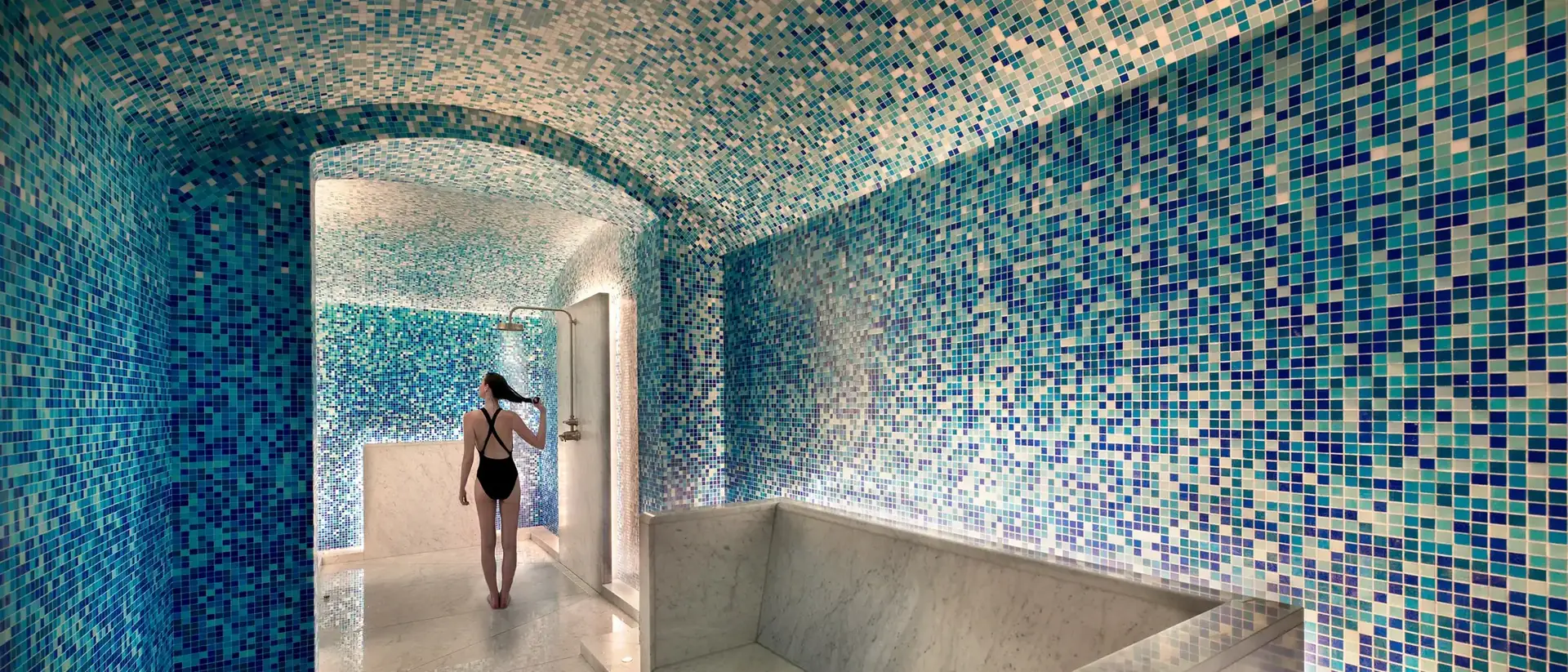 Mosaic-tiled hammam at The Newt in Somerset, with a guest standing beneath a vaulted blue and white mosaic ceiling beside a marble bench